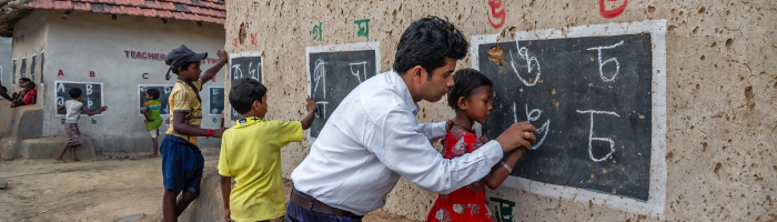 In a rural setting, a teacher passionately educates children on street using makeshift blackboards on a mud wall.